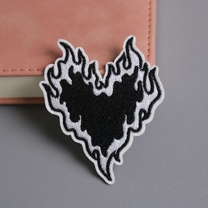 Pode incluir: Patch bordado em forma de coração preto com design de chama e borda branca. O patch está colocado em um caderno rosa claro e em uma superfície cinza. Adequado para roupas ou acessórios.