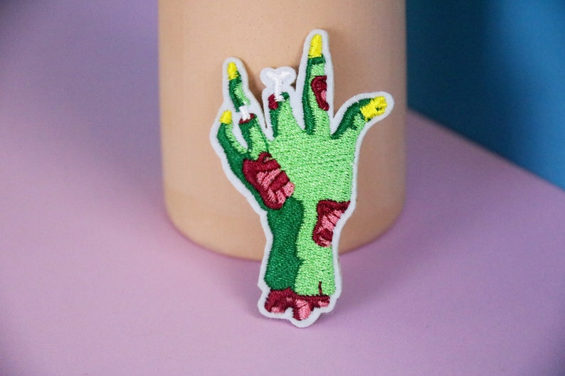 May include: Green zombie hand with red and yellow details, embroidered iron-on patch.