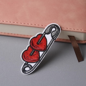 May include: White embroidered patch of a safety pin with three red hearts on it.