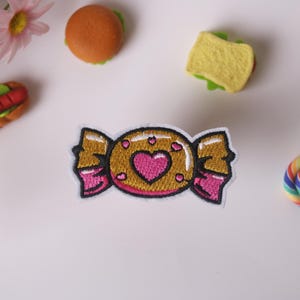 Puede incluir: Parche bordado para planchar de un caramelo rosa y dorado con un corazón en el centro. El caramelo está envuelto en un envoltorio dorado con cintas rosas.