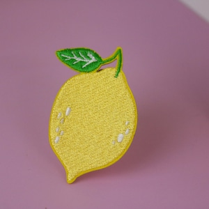 May include: An embroidered lemon with a green leaf, on a pink background.