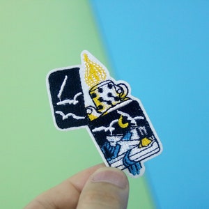 May include: A black and white embroidered patch of a lighter with a flame and a moon design. The lighter is filled with a blue and white design.