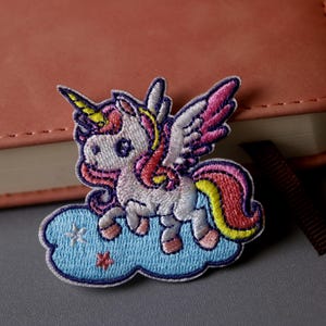 May include: A white unicorn with pink and yellow mane and tail, with wings, flying on a blue cloud with white stars. The unicorn is embroidered on a fabric patch.