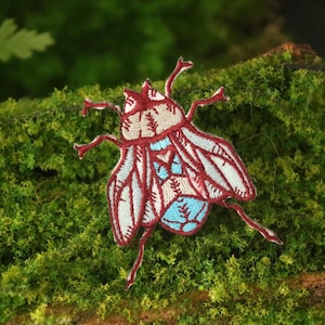 Op de afbeelding: Een geborduurde vliegenpatch in rood en wit met blauwe accenten. De patch bevindt zich op een groen mossig oppervlak.