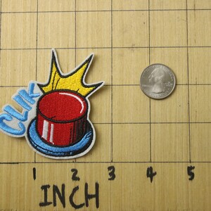 May include: A red button with a blue rim and a yellow crown on top. The button has the word "CLIK" embroidered in blue on the side. The button is about 1 inch in diameter.