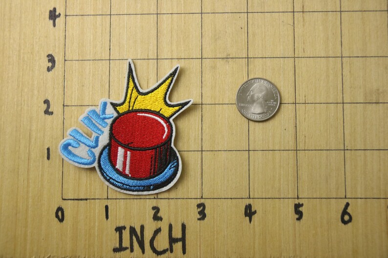 May include: A red button with a blue rim and a yellow crown on top. The button has the word "CLIK" embroidered in blue on the side. The button is about 1 inch in diameter.