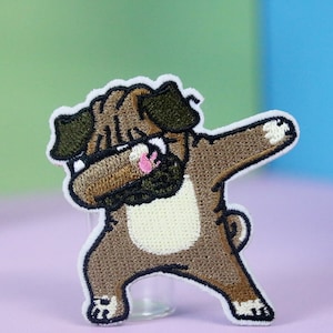 以下が含まれることがあります： ダブしている茶色と白の刺繍されたパグの犬のパッチ。パッチは白い輪郭があり、布製です。