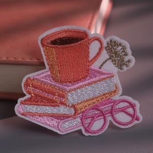May include: An embroidered patch featuring a stack of three books in shades of pink and orange, topped with a coral-coloured mug. A pair of pink round-rimmed glasses rests at the base of the books, accompanied by a small, embroidered gold flower.