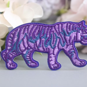 Könnte beinhalten: Gestickter lila Tiger-Aufnäher mit türkisfarbenen Akzenten. Der Tiger wird in einer Laufpose dargestellt, wobei detaillierte Stiche einen strukturierten Effekt erzeugen. Dieser Aufnäher eignet sich für Kleidung oder Accessoires.