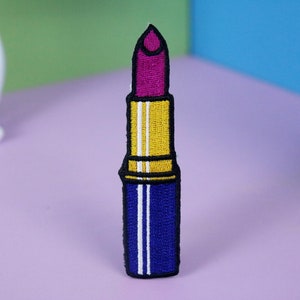 May include: A bright pink lipstick with a gold cap and a blue base. The lipstick is embroidered on a fabric patch.