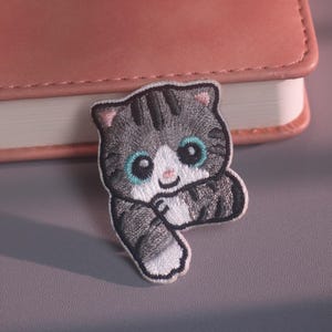 May include: An embroidered iron-on patch featuring a cartoon-style grey tabby kitten with dark grey stripes, large teal eyes, a white muzzle, and a small pink nose. The kitten is depicted with one paw raised, showcasing detailed stitching and a white border.
