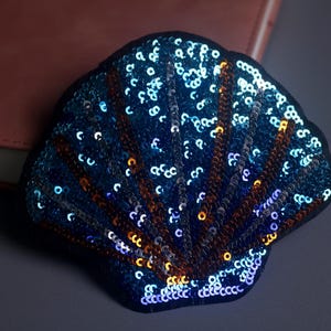 May include: A sequined seashell patch in shades of blue, with accents of gold and brown. The patch is on a dark background, with a hint of a pink notebook visible in the background. The sequins create a shimmering effect.