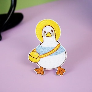 May include: Embroidered patch featuring a cartoon duck with a yellow halo, a blue shirt, and a yellow bag. The duck has brown feet and a yellow strap. The patch is white, blue, yellow, and brown. The patch is likely for clothing or accessories.