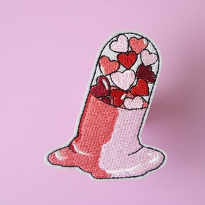 May include: Embroidered iron-on patch of a pink and red pill filled with pink and red hearts.