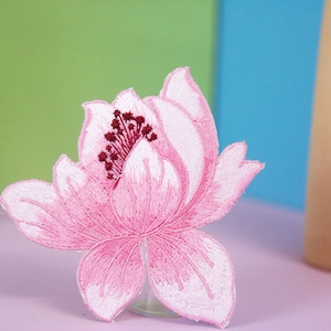 以下が含まれることがあります： ピンクの刺繍が施された花のパッチ。白い中心と濃い赤色の雄しべがあります。パッチはピンクの表面にあります。