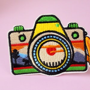 Może przedstawiać: Kolorowa haftowana naszywka do prasowania przedstawiająca aparat fotograficzny z widokiem zachodu słońca w wizjerze. Aparat jest żółty, zielony, niebieski, czerwony i pomarańczowy.
