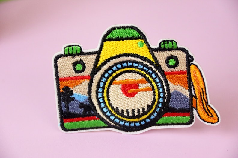 Puede incluir: Un parche bordado de colores para planchar que representa una c&aacute;mara con una escena de puesta de sol en el visor. La c&aacute;mara es amarilla, verde, azul, roja y naranja.
