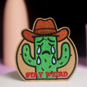 Könnte beinhalten: Gestickter Aufnäher mit einem grünen Kaktus, der einen braunen Cowboyhut trägt und blaue Tränen weint. Der Aufnäher trägt den roten Schriftzug "STAY WEIRD". Der Aufnäher ist beige umrandet.