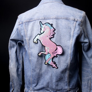 May include: Pale blue denim jacket featuring a large sequined unicorn patch on the back. The unicorn is pink with blue and light blue details. The jacket has a classic design with button fastenings and pockets.