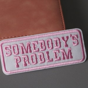 May include: White rectangular patch with pink embroidered text that reads "Somebody's Problem".