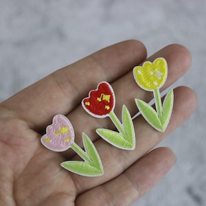 Puede incluir: Tres parches bordados para planchar en forma de tulipanes, en rosa, rojo y amarillo. Cada parche tiene un tallo y hojas verdes.