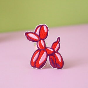 May include: Embroidered patch of a red balloon dog with pink and purple detailing. The patch has a white border and is set against a pink and green background. This patch is suitable for decorating clothing or accessories.