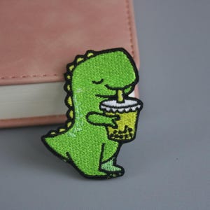May include: Embroidered patch showing a green dinosaur drinking bubble tea. The dinosaur has a happy expression and is holding the drink. The patch has a black border and is suitable for clothing or accessories. The patch is approximately 5 cm in height.