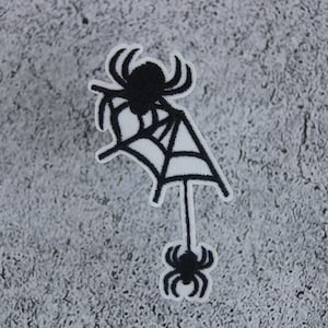 Pode incluir: Etiqueta bordada preta de ferro de uma aranha em uma teia com uma aranha menor pendurada na parte inferior.