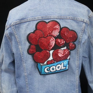 May include: A light blue denim jacket with a red sequined heart patch on the back. The patch features a box with the word "COOL" in blue and white letters.