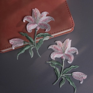 Peut inclure: Écussons brodés de fleurs de lys roses et blanches avec des feuilles vertes. Les écussons sont placés sur une surface gris foncé à côté d'un carnet en cuir marron. Les écussons sont détaillés et délicats.