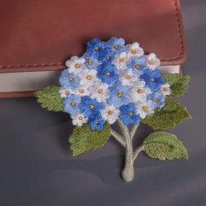 May include: Embroidered hydrangea flower patch with blue and white petals, green leaves, and a gray stem. The floral design is detailed and sits on a gray surface next to a pink notebook.