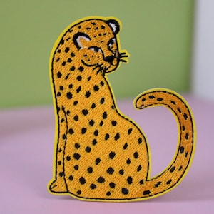 Può includere: Un toppa ricamata di colore giallo da stirare con un ghepardo seduto con la coda arrotolata attorno al suo corpo. Il ghepardo ha macchie nere.