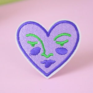 Pode incluir: Etiqueta bordada em forma de coração roxo com um rosto estilizado em fio verde e roxo.