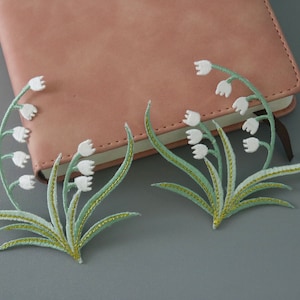 Peut inclure: Deux patchs brodés de fleurs de muguet blanches avec des feuilles vertes et des accents dorés.
