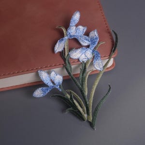 May include: Embroidered iris flower patch with blue and white petals and green stems. The floral design is placed next to a closed, brown leather-bound notebook on a gray surface. The patch is a decorative element.