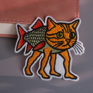 以下が含まれることがあります： 魚の体と足を持つ猫の刺繍パッチ。猫はオレンジ色の毛皮に黒い縞模様、緑色の目、白いひげがあります。魚の尾は赤く、体は緑色です。パッチには白い縁取りがあります。