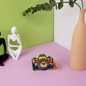 Puede incluir: Un parche bordado de colores para planchar con forma de c&aacute;mara vintage con lente amarilla, detalles en verde y azul y una correa marr&oacute;n. El parche est&aacute; sobre una superficie rosa.