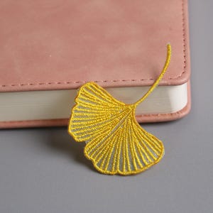 Könnte beinhalten: Ein gelb gesticktes Ginkgoblatt, das auf einer grauen Oberfläche neben einem rosa ledergebundenen Notizbuch liegt. Das filigrane Design des Blattes und seine leuchtende Farbe kontrastieren mit der weichen Textur des Notizbuchs.