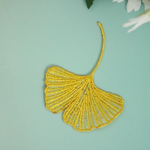 May include: A yellow ginkgo leaf-shaped applique, featuring intricate stitching details. The applique is set against a light teal background, with white flowers in the periphery. This decorative element is suitable for various craft projects.