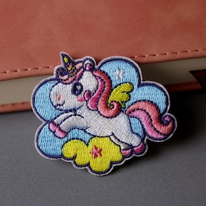 May include: A white unicorn with a pink mane and tail, wearing a yellow star on its head, is flying on a blue cloud with a yellow star. The unicorn is embroidered on a patch.
