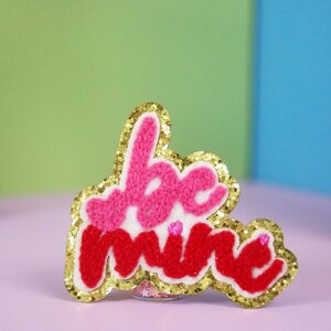 Puede incluir: Un parche con ribete dorado con las palabras "be mine" en letras peludas de color rosa y rojo.