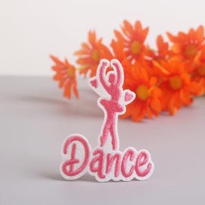 Op de afbeelding: Een roze en witte geborduurde patch met een ballerina en het woord "Dance". De ballerina staat in een danshouding met de armen omhoog. De patch staat tegen een vage achtergrond van oranje bloemen.