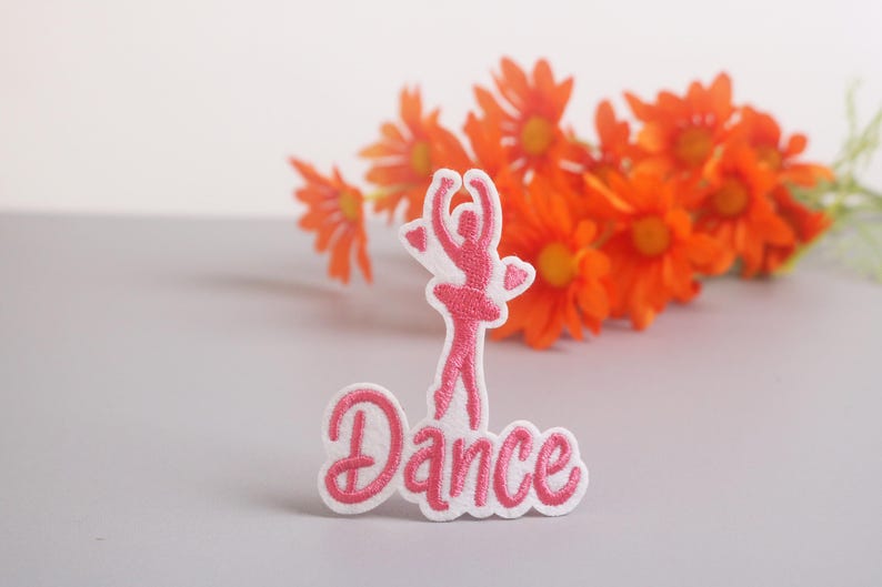 Op de afbeelding: Een roze en witte geborduurde patch met een ballerina en het woord "Dance". De ballerina staat in een danshouding met de armen omhoog. De patch staat tegen een vage achtergrond van oranje bloemen.