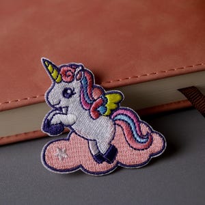May include: An embroidered patch featuring a white unicorn with a pink, blue and yellow mane and tail, standing on its hind legs on a pink cloud. The unicorn has a yellow horn and is set against a white background.