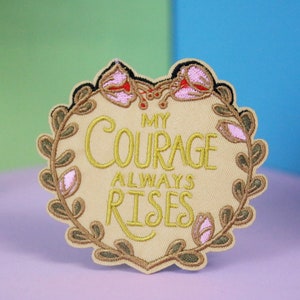 Puede incluir: Un parche bordado de color beige en forma de corazón con una corona floral dorada y el texto "My Courage Always Rises".