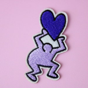 May include: Embroidered patch featuring a stylized figure in lavender holding a deep purple heart. The figure has a black outline and is on a white background. The patch is set against a light pink background.