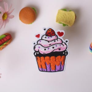 May include: An embroidered iron-on patch of a pink and white cupcake with a red cherry on top. The cupcake is decorated with white sprinkles and has a dark brown chocolate base. The cupcake is sitting on a purple and orange base.