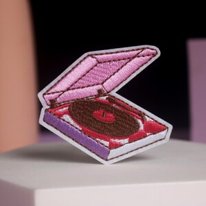 May include: Embroidered patch of a pink and purple record player with a brown record. The patch is open, showing the record inside. The patch is on a white surface.