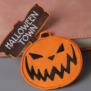 May include: Two embroidered iron-on patches. One is brown with white text that says "Halloween Town". The other is orange and depicts a jack-o'-lantern with a black face.
