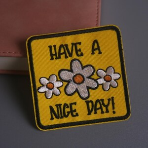 Peut inclure: Écusson brodé jaune avec bordure noire, portant l'inscription "HAVE A NICE DAY !" et trois fleurs marguerites. Les fleurs ont des pétales blancs et des centres orange. L'écusson est de forme carrée.